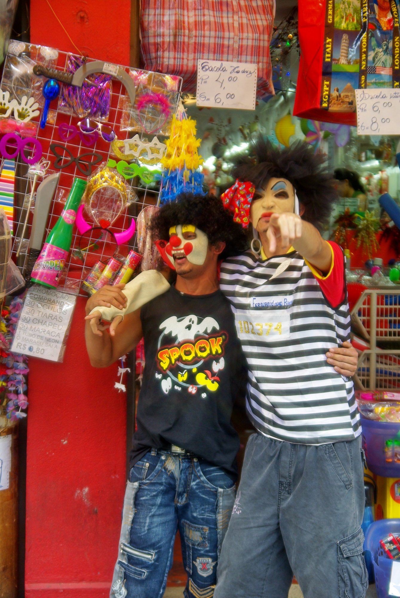Dois rapazes fantasiados de palhaço posam em frente a loja de artigos de festa com óculos e tiaras coloridos