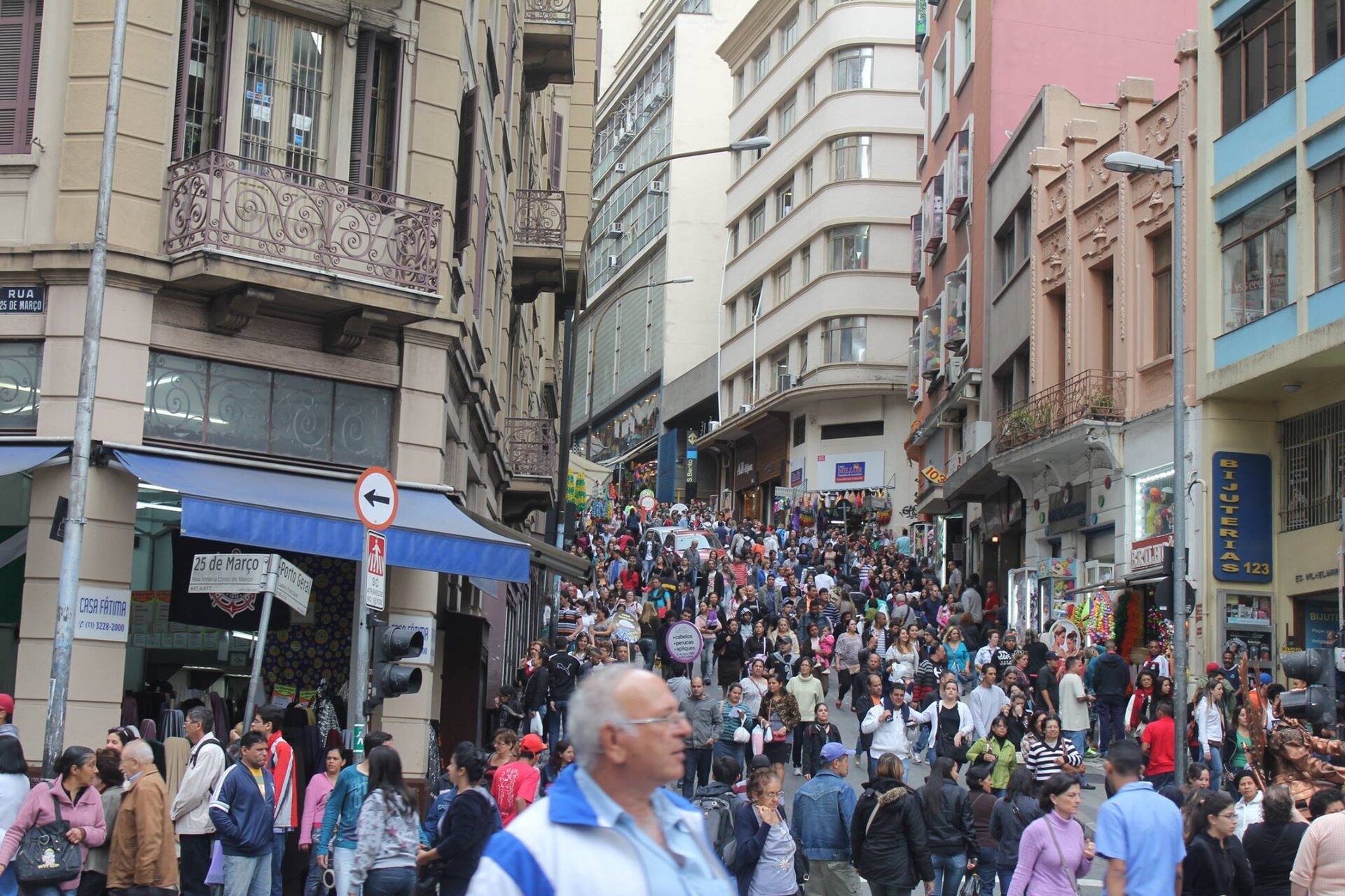 Rua 25 de Março cheia de pedestres entre prédios antigos e lojas com toldos